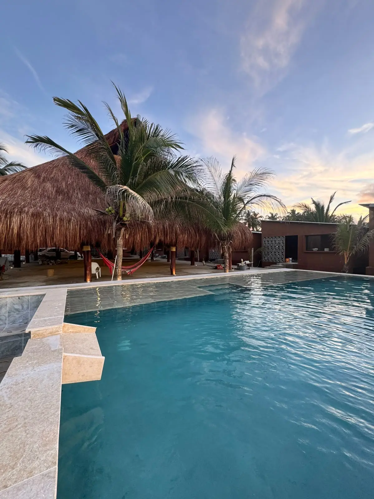 Piscina de Mokano Beach Hotel con borde en piedra clara y palapa al fondo